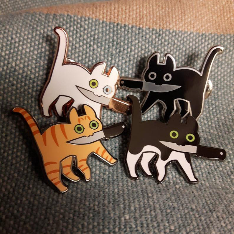 Group of different knifecat enamel pins together on a grey blanket.