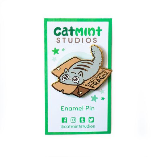 The Emotionally Fragile Pin on its white backing card. “Catmint Studios” is across the top in green. Below the pin, it says “Enamel Pin” and “@catmintstudios” with the icons representing facebook, instagram, tumblr, and twitter.