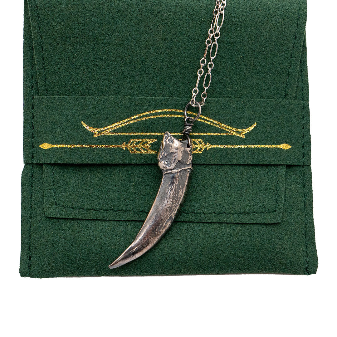 the silver badger claw pendant resting against a green jewelry pouch. the pouch has a golden bow and arrow design in the center.