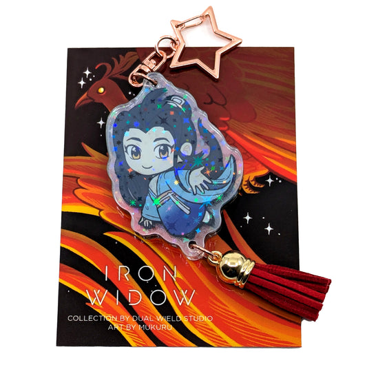 A product photo of Side 1 of the Yizhi Holographic Charm. This side illustrates Yizhi wearing blue traditional clothing, reaching out toward the viewer. The charm has a gold star shaped clasp, a red tassel, and the acrylic has a holographic effect. The charm is attached to its backing card, which has an illustration of a red feathery phoenix with a glowing yellow eye.
