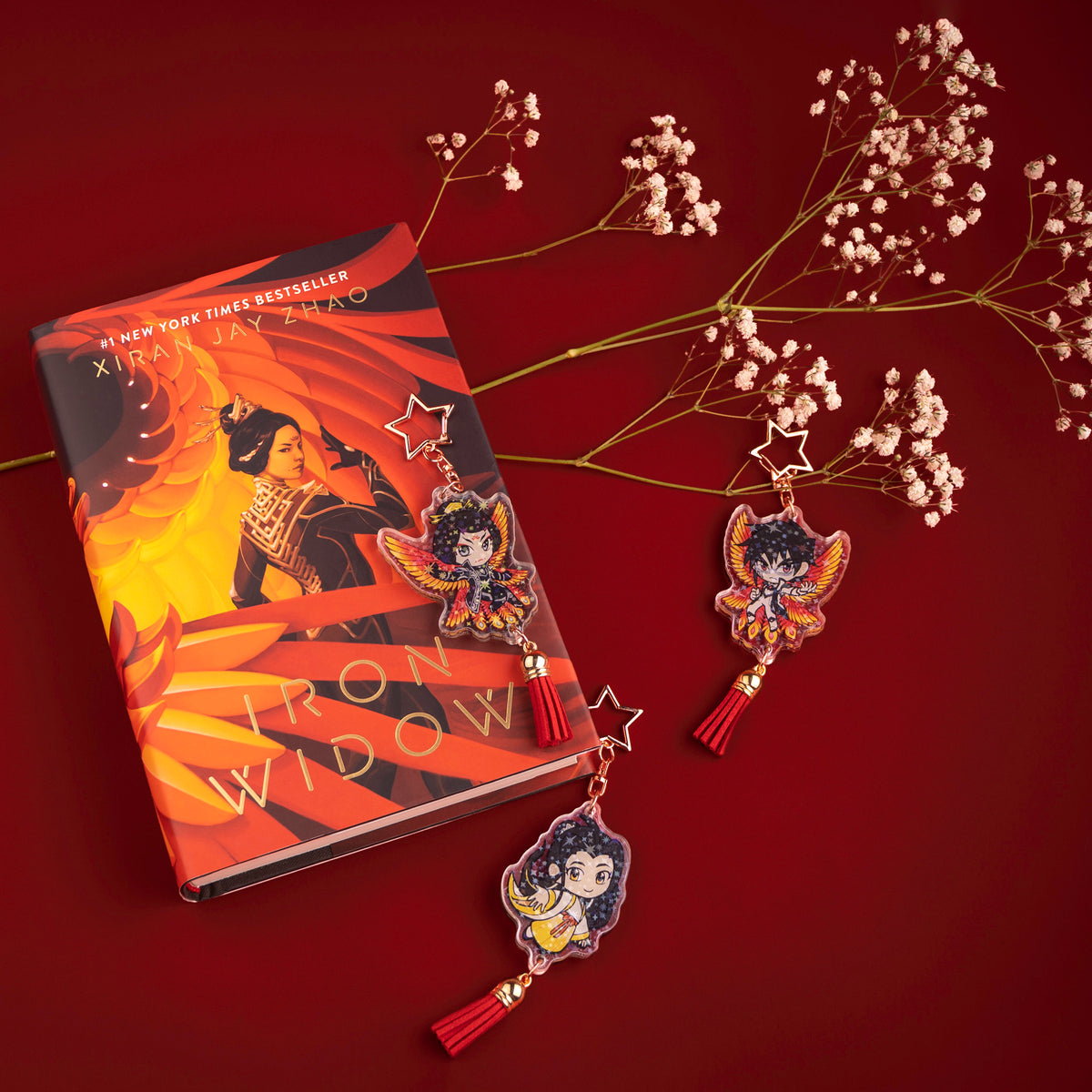 The Zetian, Yizhi, and Shimin holographic charms, all showing their Side 1, respectively, are scattered around a hardback copy of Iron Widow, accompanied by a baby’s breath flower stem.