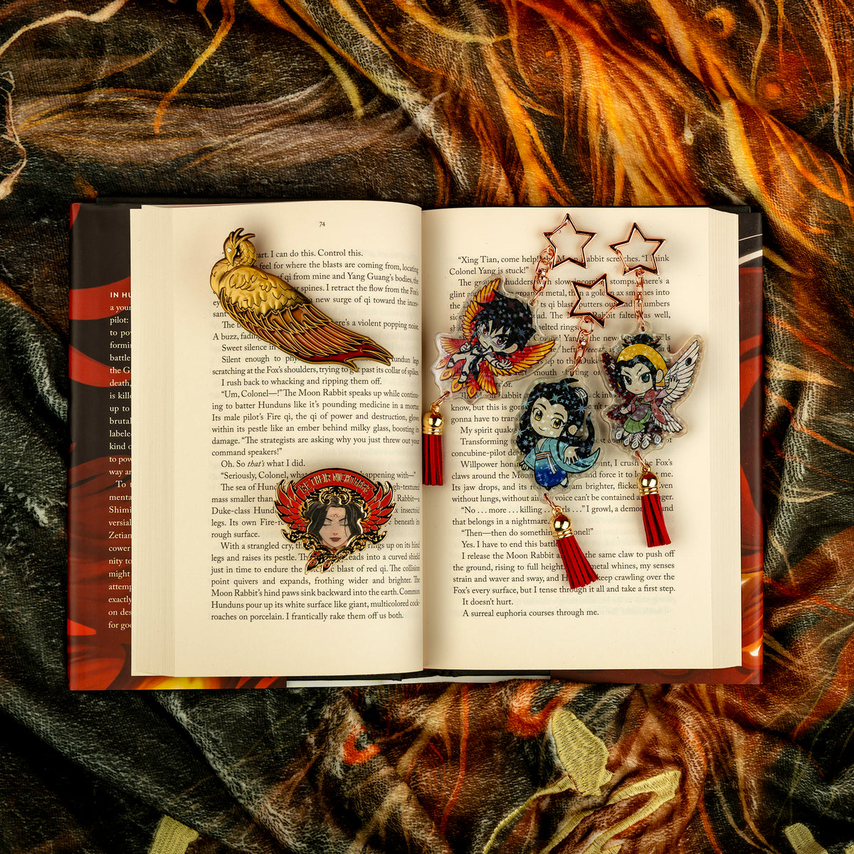 The Vermillion Bird Hair Clip, the Be Their Nightmare Pin, and all three Holographic charms (Xetian, Shimin, and Yizhi) are positioned over a flat-laid open hardback copy of Iron Widow. The book itself is cozied into the Phoenix Usurping Dragon Blanket. All of the items’ golden aspects shine against the pages.