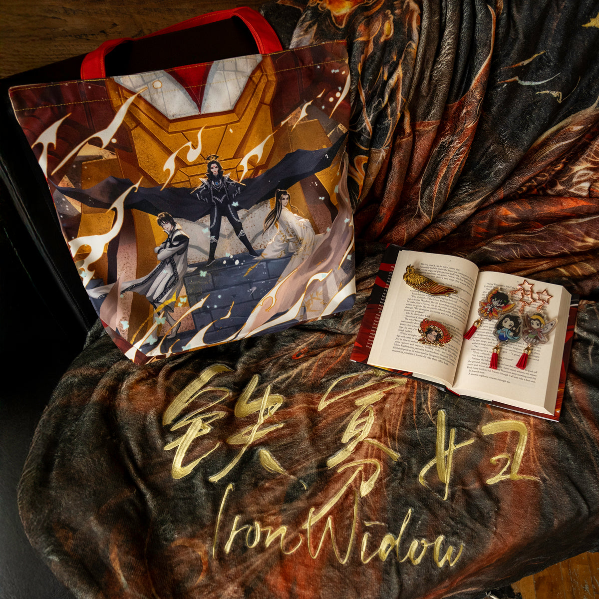 The Phoenix Usurping Dragon Blanket is draped over a sofa. On top of it rests the Iron Triangle tote, the trio side facing forward. An open copy of Iron Widow lays beside it, with the Vermillion Bird Hair Clip, the Be Their Nightmare Pin, and all three Holographic charms (Xetian, Shimin, and Yizhi) laying on top of the pages.