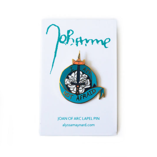 A product photo of the I Am Not Afraid Pin on its white backing card. Joan of Arc’s signature is across the top in light teal. Below the pin it reads “Joan of Arc Lapel Pin” and “alyssamaynard.com”