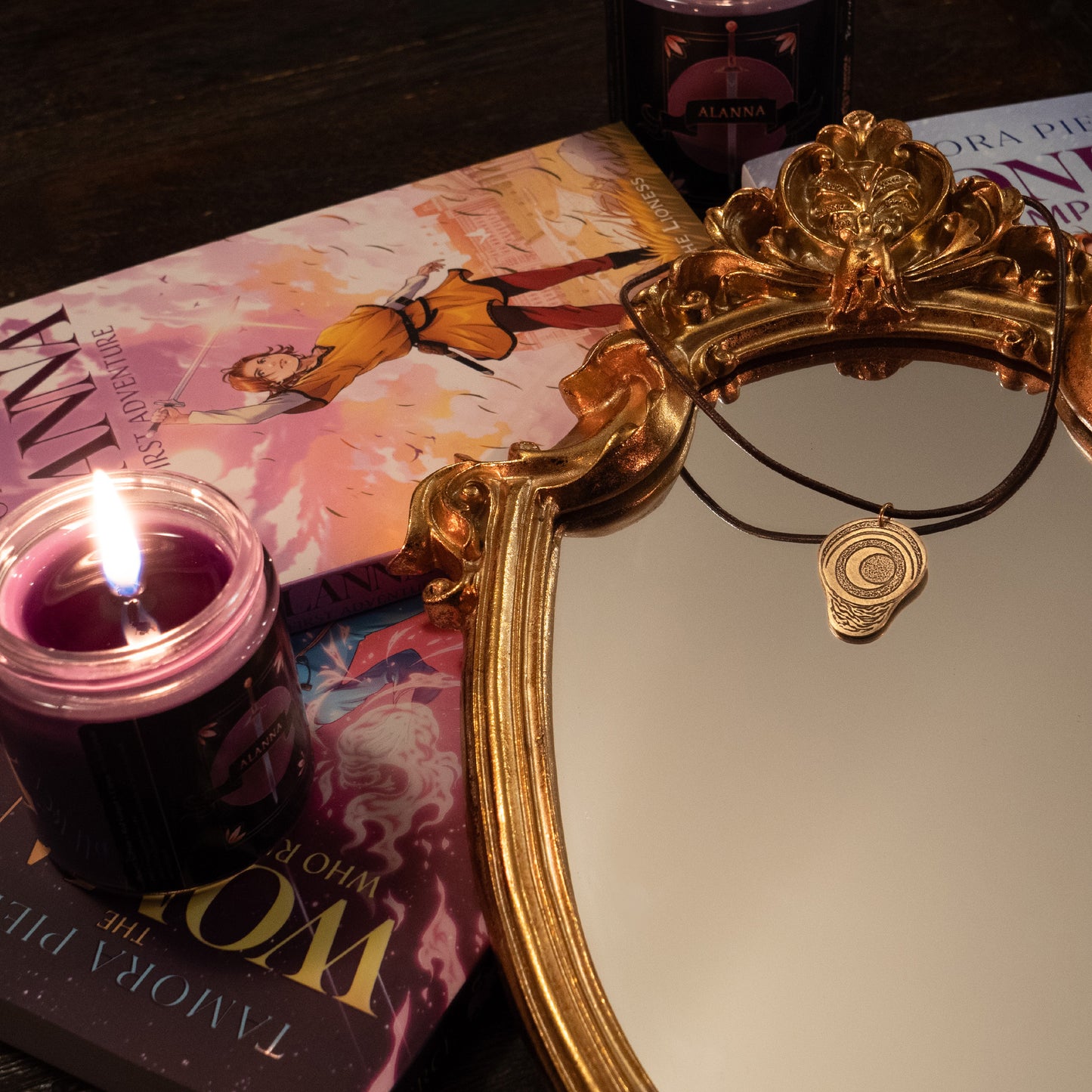 A photo of the necklace laying on a gilded mirror, surrounded by Tamora Pierce books and Tamora Pierce candles by Briarwick.