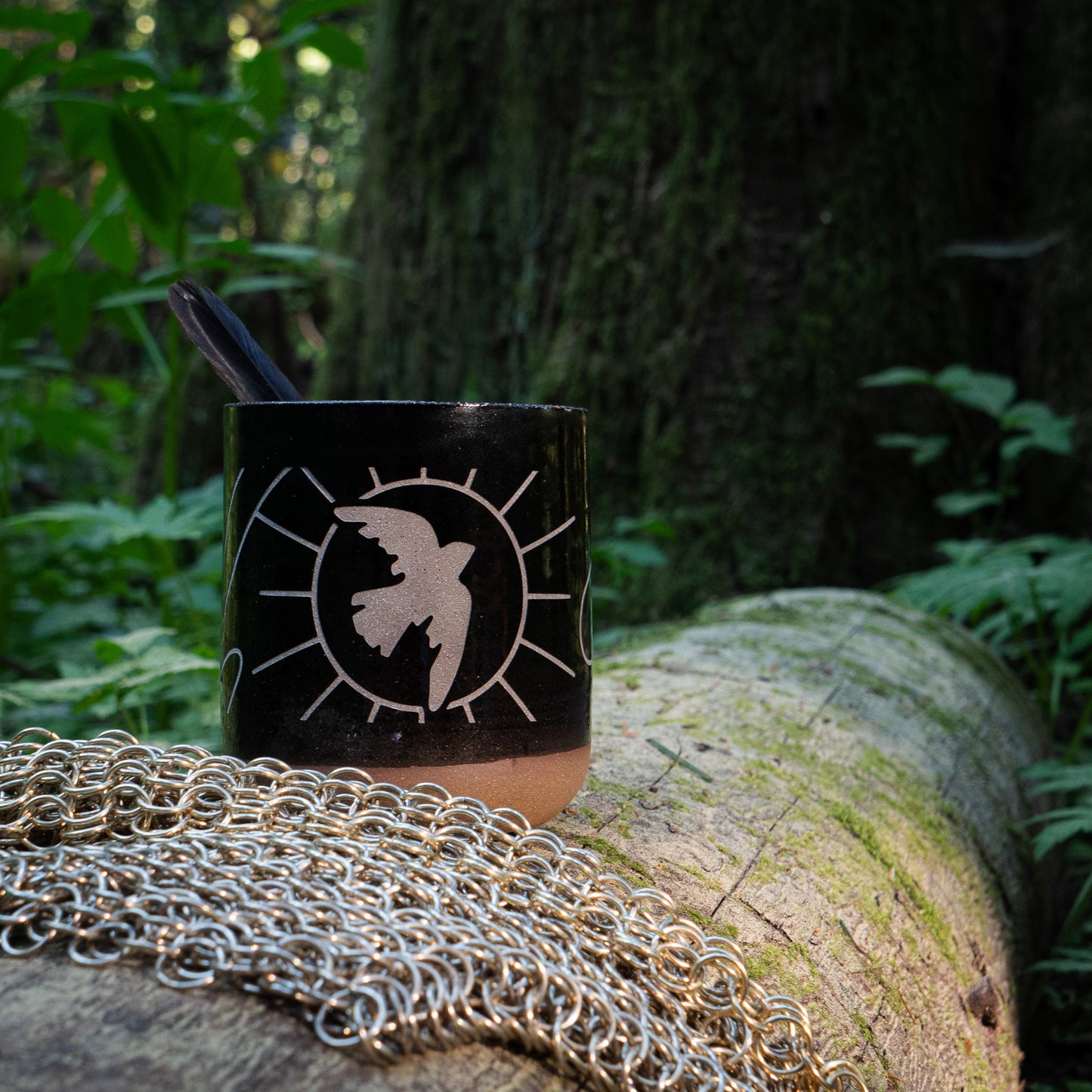 A photo of the dancing dove mug beside chainmail on a log in the forest, a feather poking out from inside the mug.