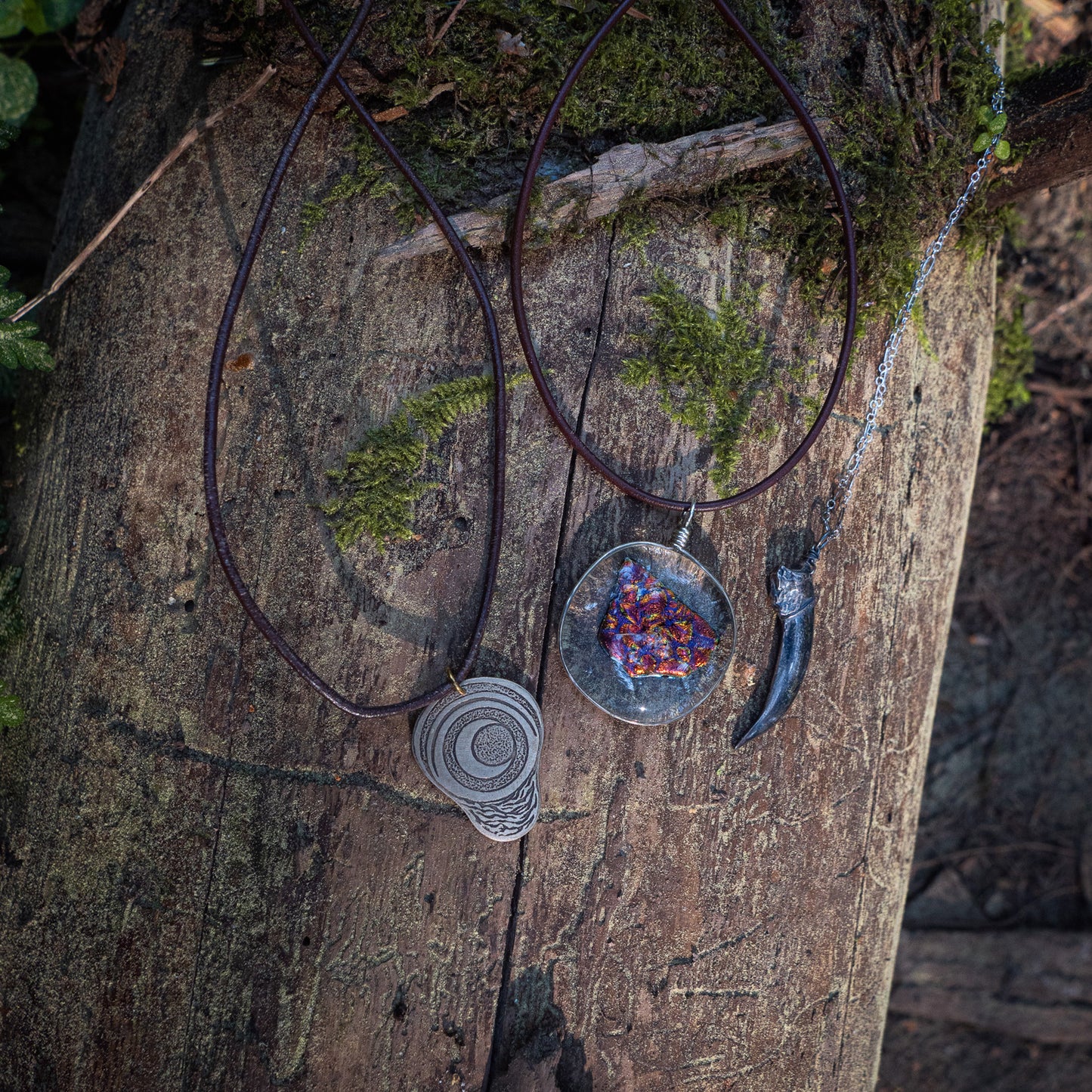 The pregnancy charm, ember glass necklace, and silver badgerclaw necklace laying beside one anther on a mossy log