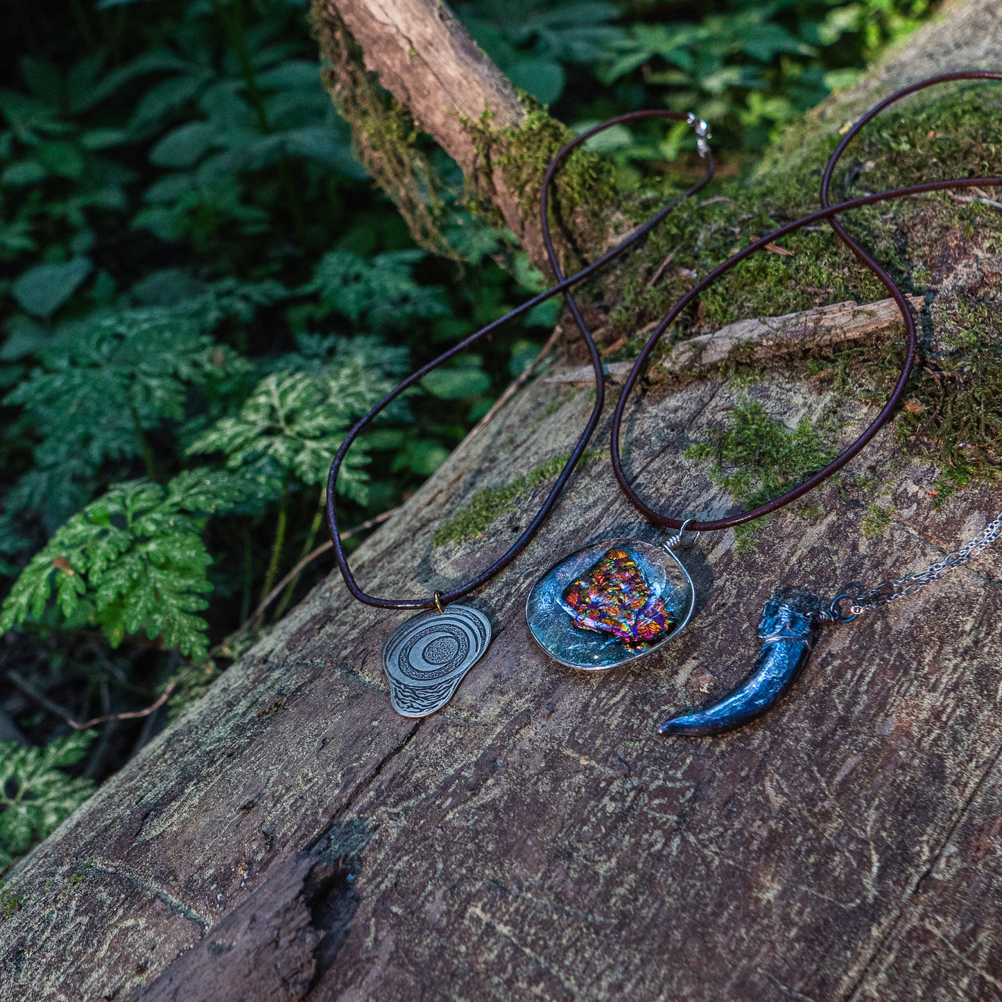 The pregnancy charm, ember glass necklace, and silver badgerclaw necklace laying beside one anther on a mossy log amidst ferns.
