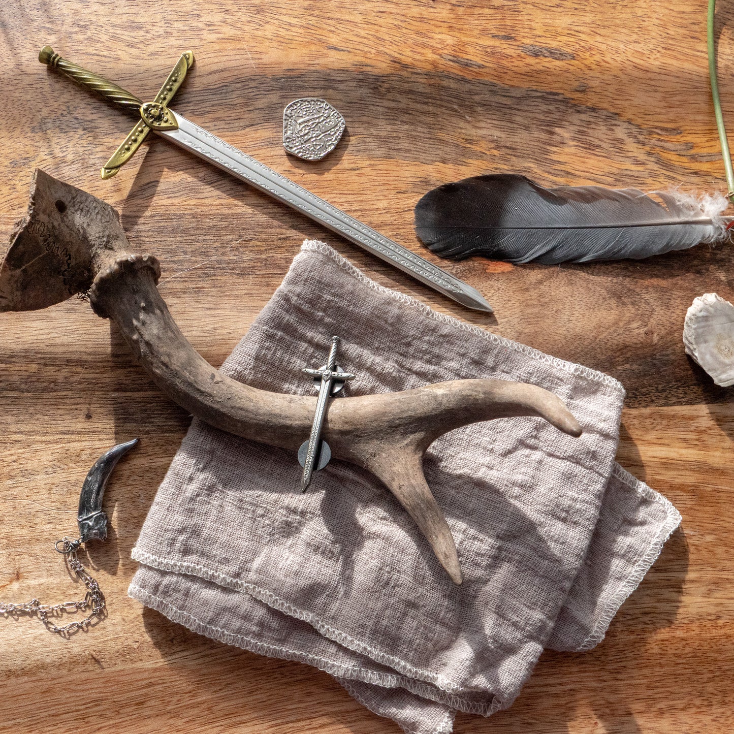 A photo of the Tamora Pierce: Lightning Mini sword arranged on a table with the Lightning Pin, the Badger Claw Necklace, and assorted objects such as a black feather and a deer antler.