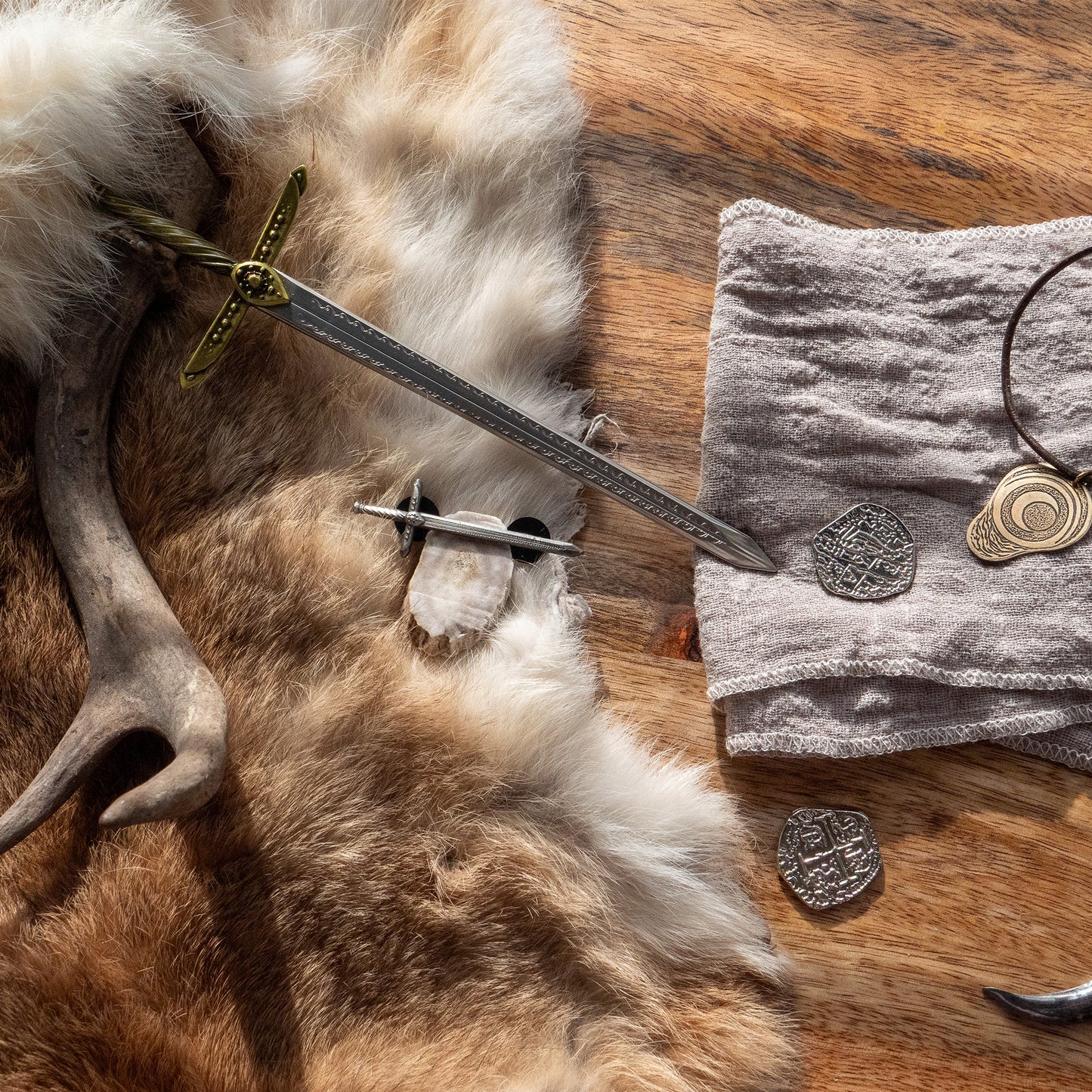 A photo of the Tamora Pierce: Lightning Pin arranged on a table with the Lightning Mini Sword, the Badger Claw Necklace, and assorted objects such as a black feather and a deer antler.