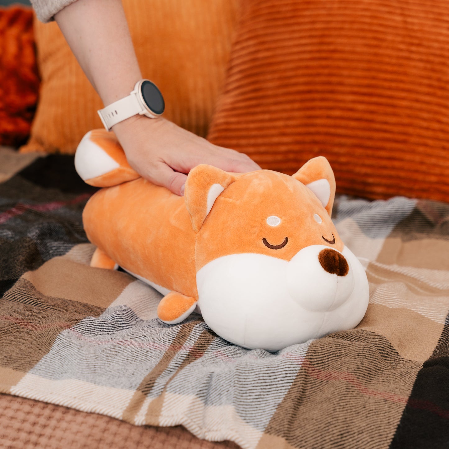 The Shiba cushion sits on a couch, with a model's hand resting on its back.