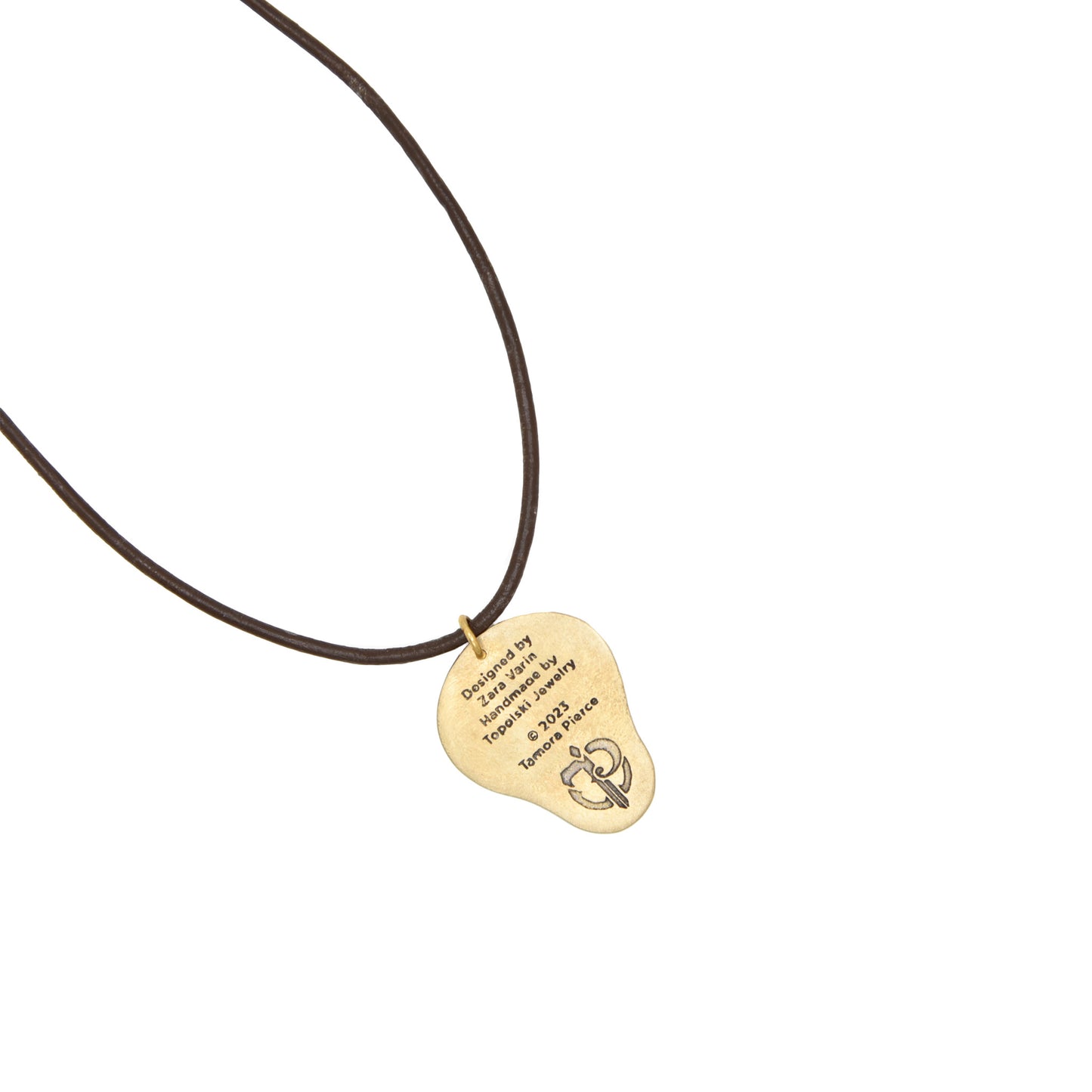 A product photo of the back of the brass pregnancy charm. Etched onto the back are credits: "Designed by Zara Varin, Handmade by Topolski Jewelry, Copyright 2023 Tamora Pierce." The Tamora Pierce sigil is etched below the credits.
