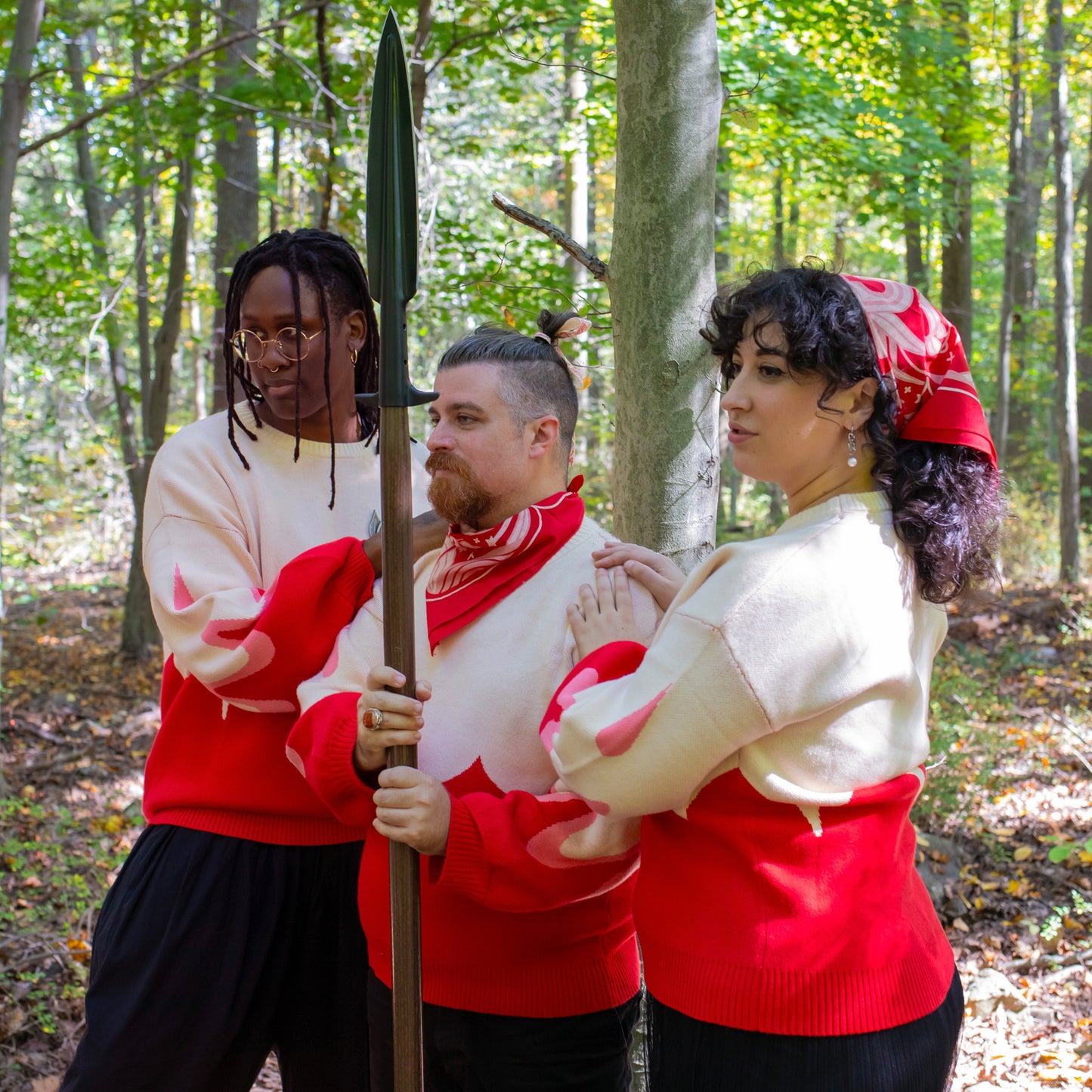 Three models wearing the Sacred Blood Sweater are standing side by side in the forest. The central model carries a spear and the models to the side of him have their hands upon his shoulders. The sweater is shown paired with the Sacred Blood bandana, worn around the neck of the central model and around the hair of the model to the right.