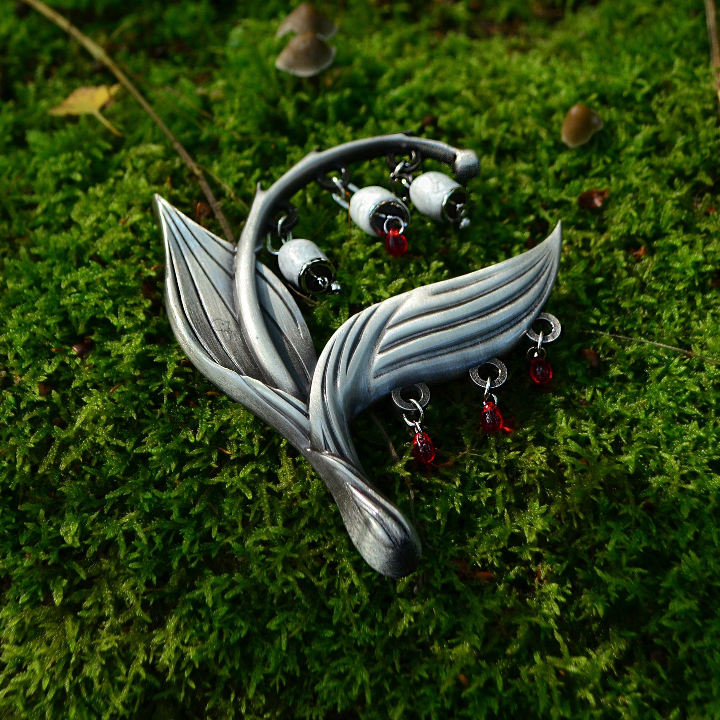 The Sacred Blood Pin is pictured laying on vibrant green moss that brilliantly contrasts the red on its beads.