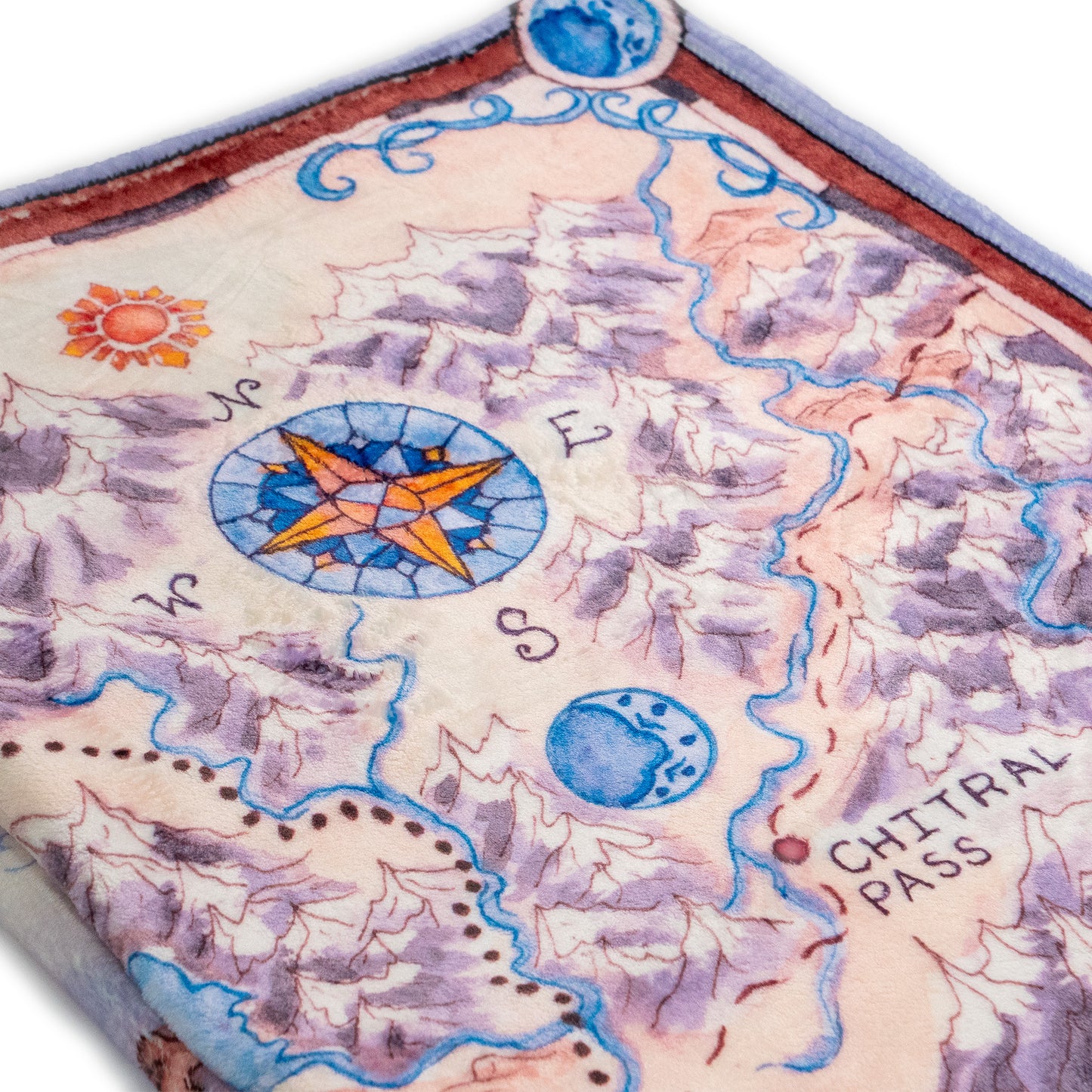 A closeup of the Tortall Map Blanket, focusing on the compass rose, which has an orange sun north of it, and a blue moon south of the rose. The compass rose is placed seamlessly in between mountains and rivers.