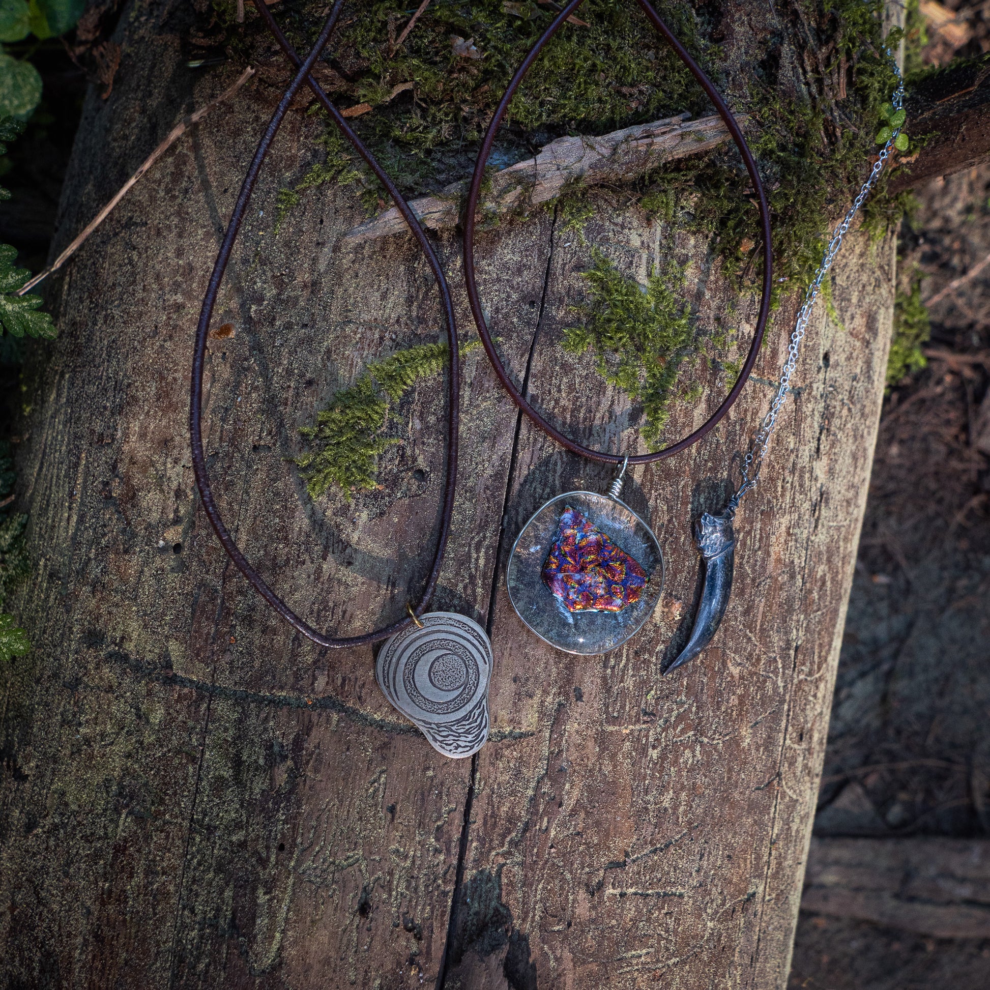 three necklaces - the badger claw, the ember glass, and the pregnancy charm - rest against tree bark and foliage.