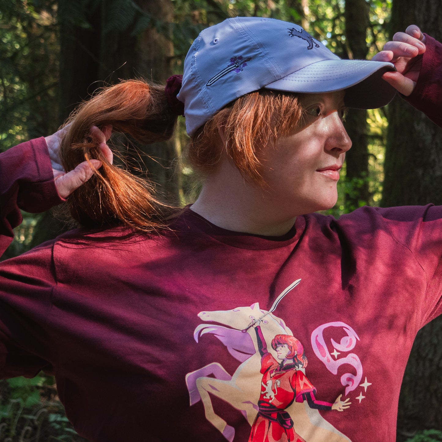 a model wears the maroon t-shirt and a lavender ballcap with a cat on it.