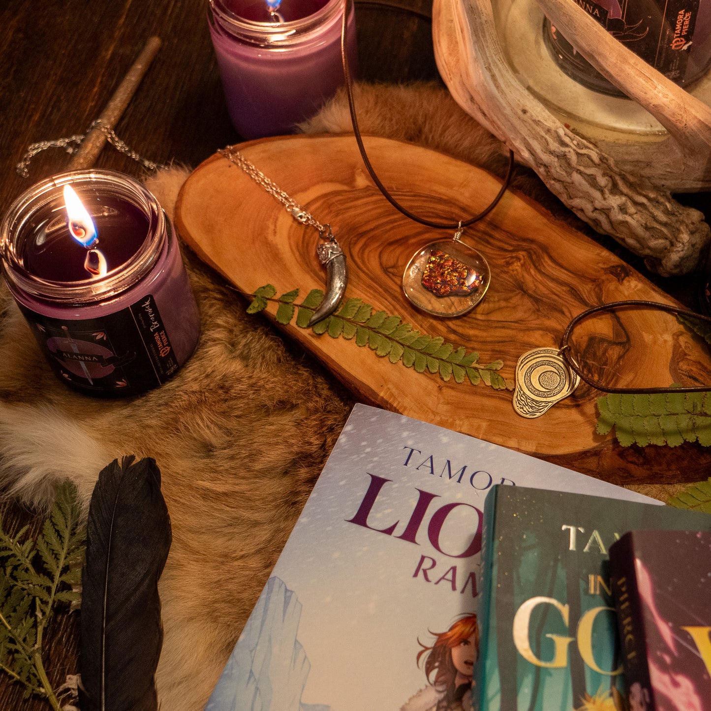 three necklaces - the badger claw, the ember glass, and the pregnancy charm - are arranged on a wooden tray with decoartive srroundings of candles, Alanna books, and foliage.