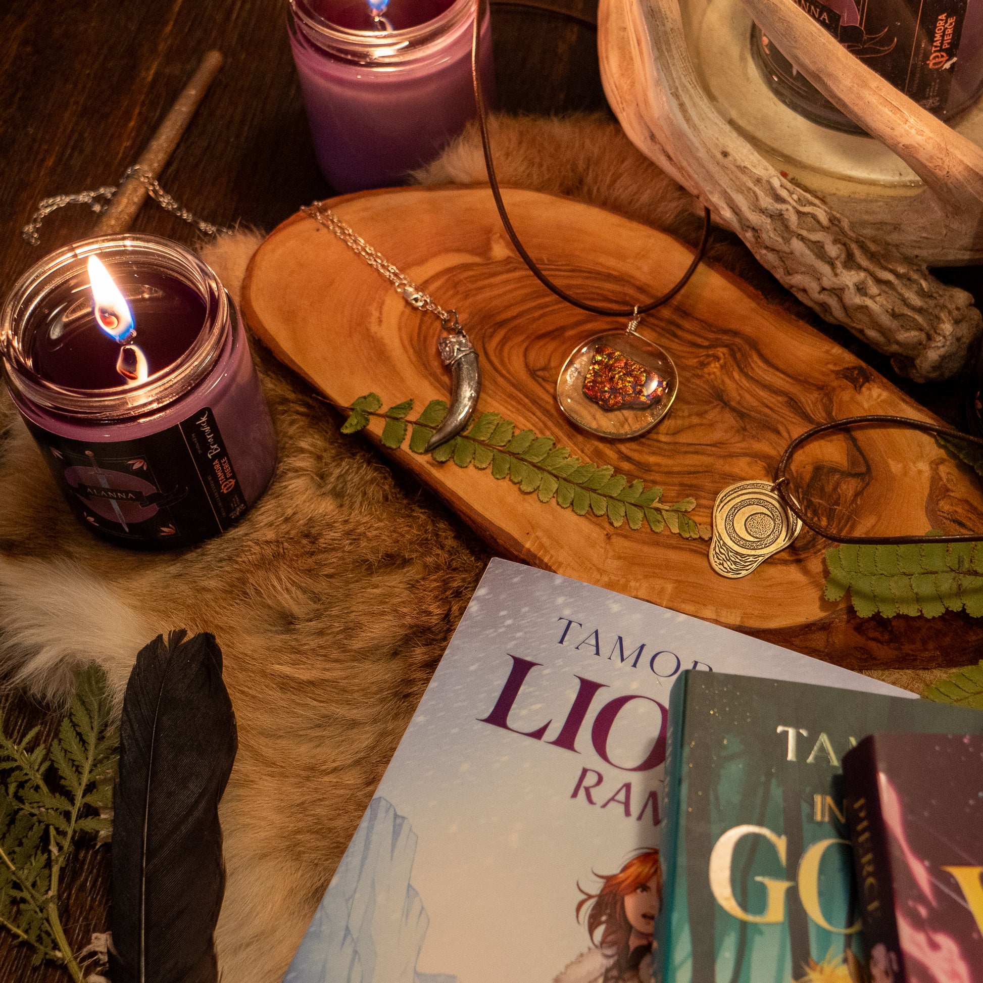 three necklaces - the badger claw, the ember glass, and the pregnancy charm - are arranged on a wooden tray with decoartive srroundings of candles, Alanna books, and foliage.