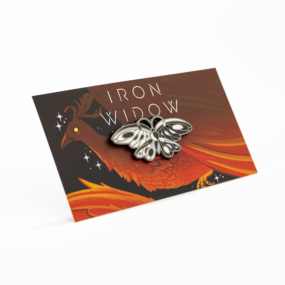 Angled view of Sparkling black and white butterfly enamel pin on red phoenix backing card with Iron Widow logo.