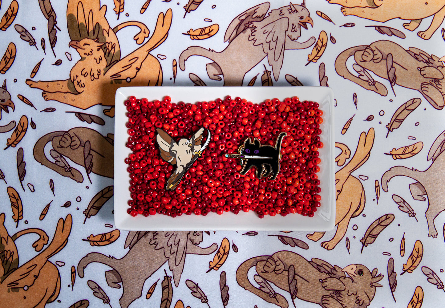 Tamora Pierce Faithful with Sword pin and Keladry Crown Sparrow pin together in a white bowl filled with red beads on Tamora Pierce: Griffin Scarf background.