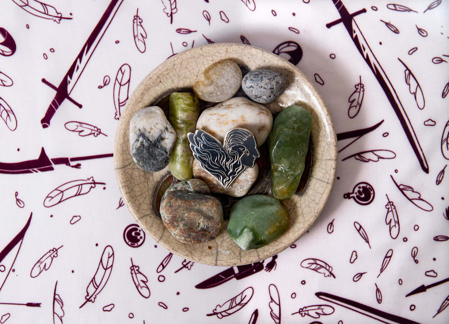 The Numair pin rests in the center of a ceramic bowl of different colored stones on a white Weapons & Spells print scarf background.
