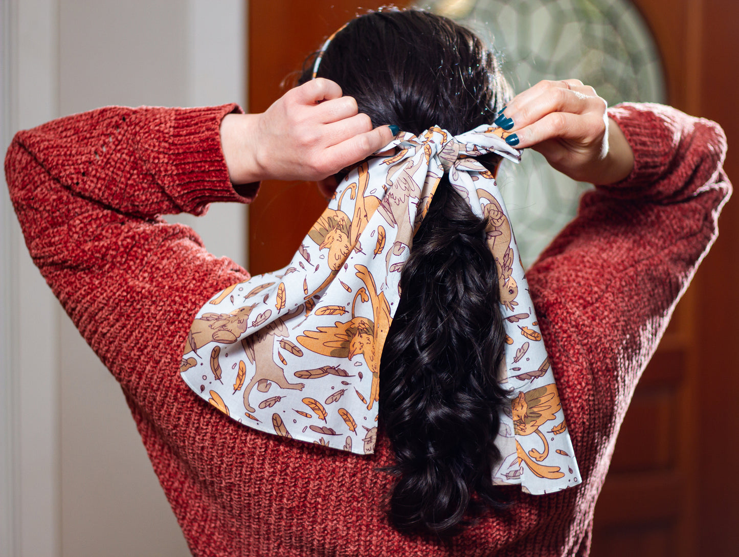 A model in a red sweater and teal nails tightens the Tamora Pierce: Griffins Scarf around her pony tail.