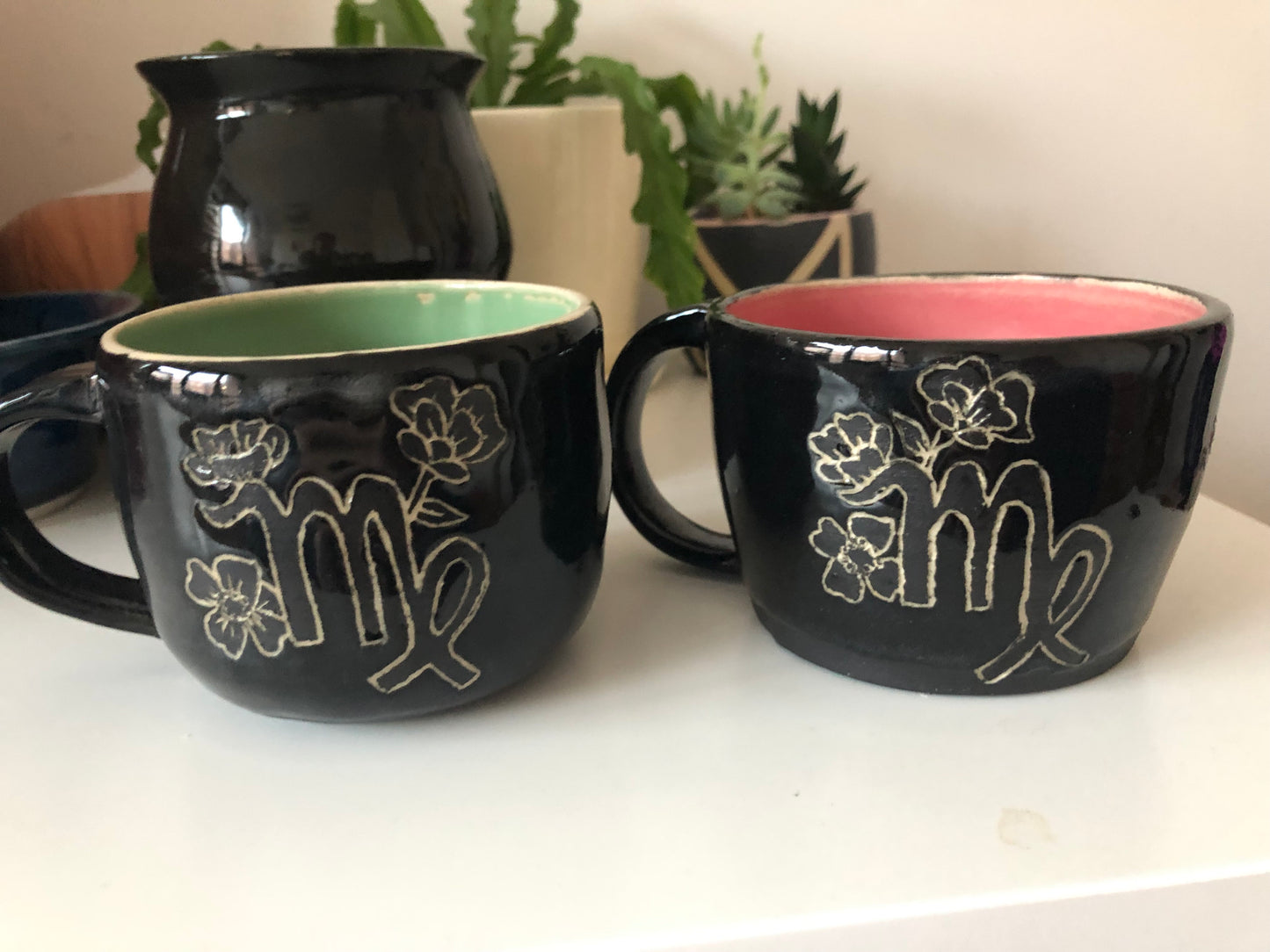 Black Virgo mugs with mint green and pink inside washes sit on a white coffee table. Mugs have Virgo symbol on them surrounded by sprouting flowers.