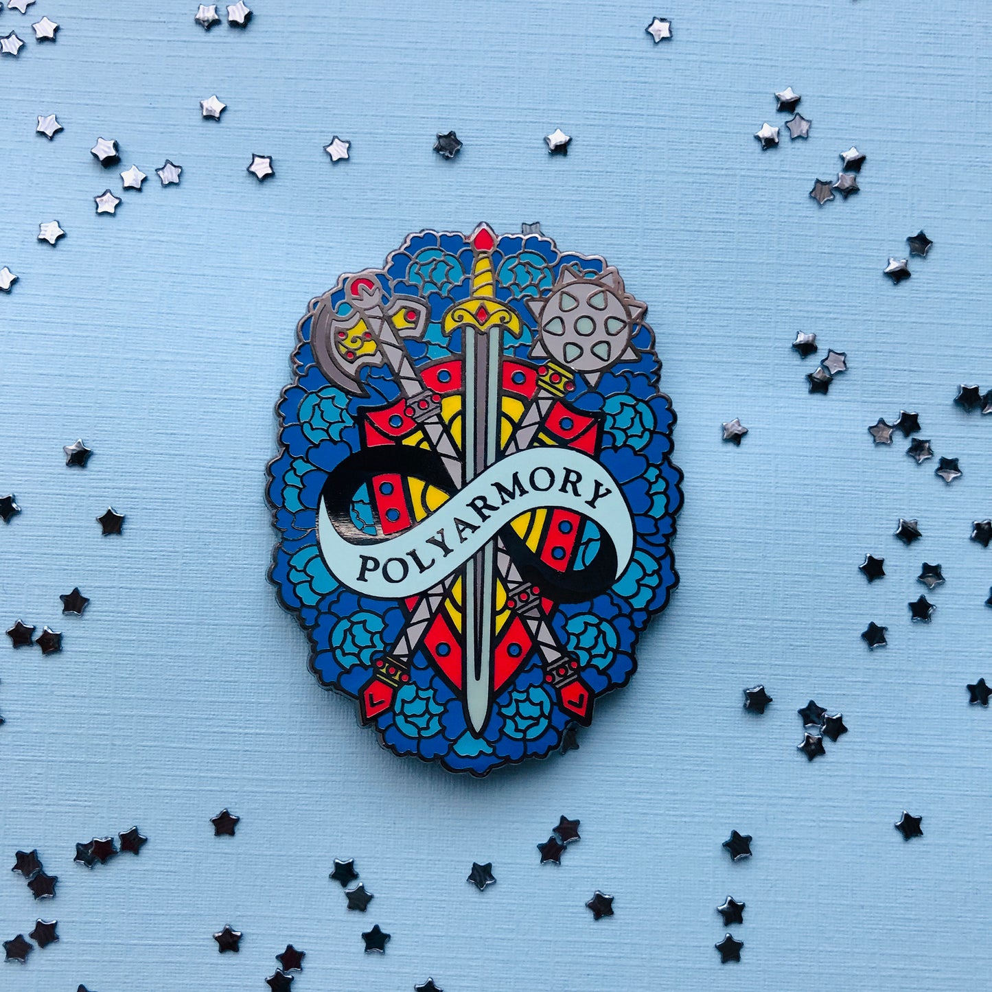 The goldcast pin features a crossed poleaxe, sword, and mace on a red and yellow shield. They all rest on a bed of roses in the different blue hues of the poly flag. A ribbon trails across the front and reads "Polyarmory."