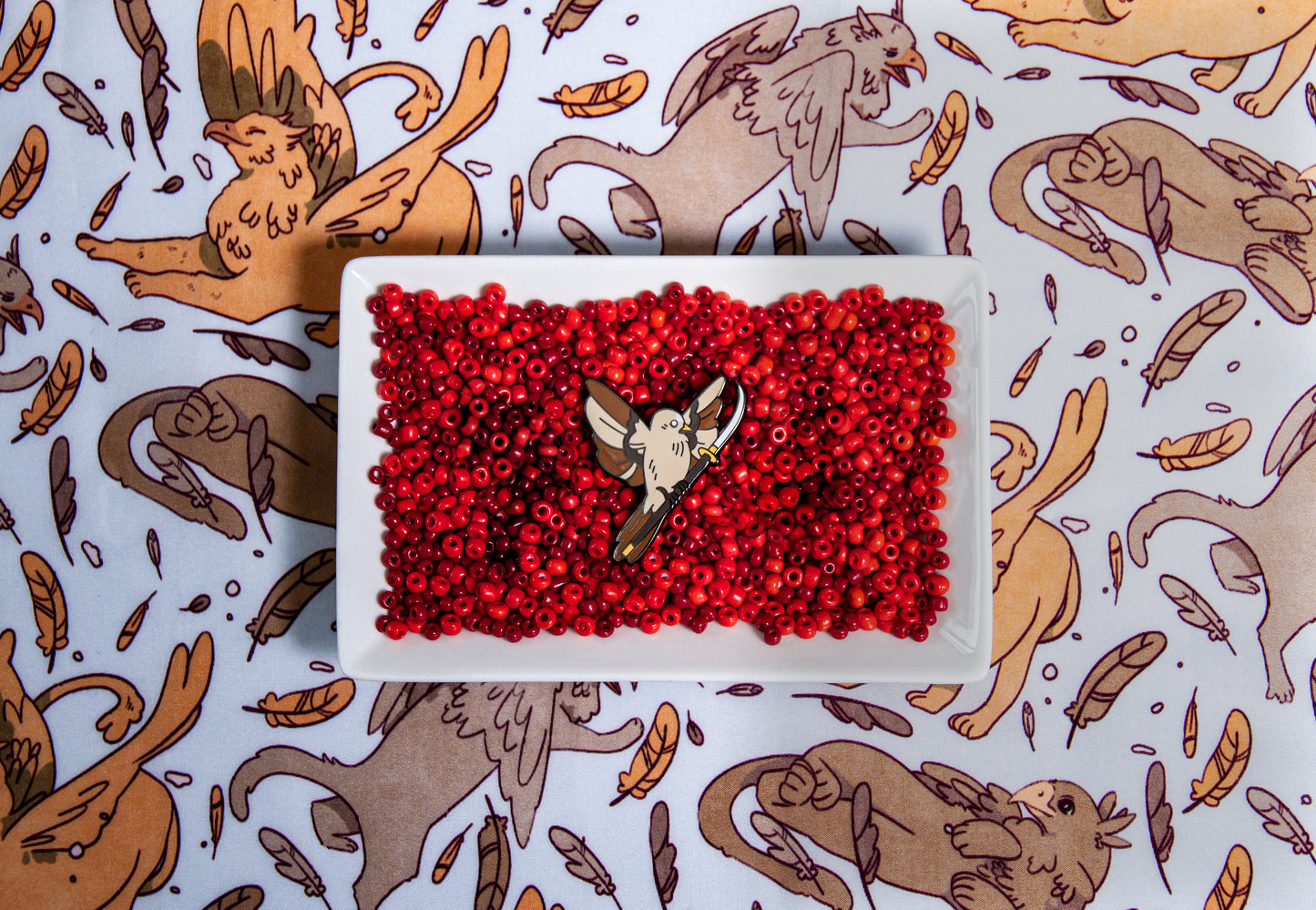 Tamora Pierce Keladry Crown Sparrow pin in a white bowl filled with red beads on Tamora Pierce: Griffin Scarf background.