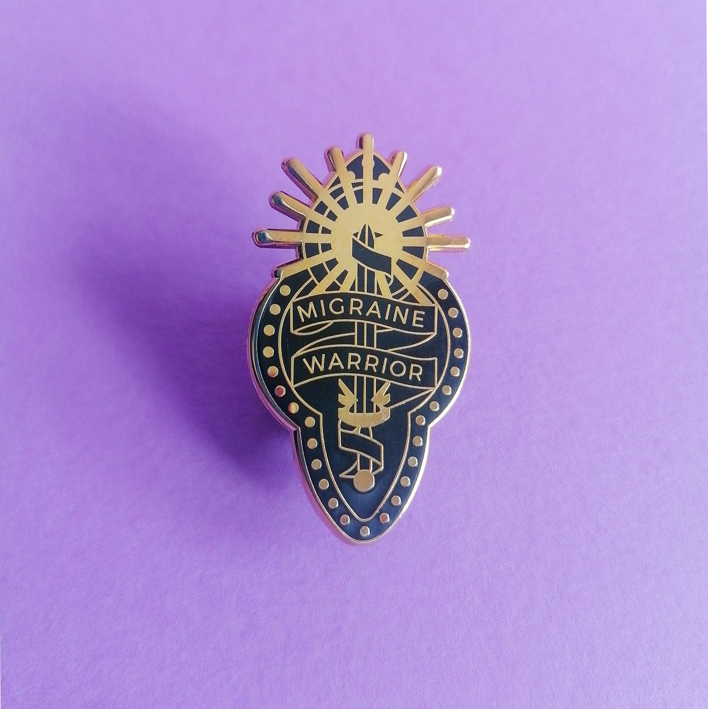 A black and gold enamel pin with design depicting a ribbon wrapped around a sword and sun, reading "Migraine Warrior" on purple background.