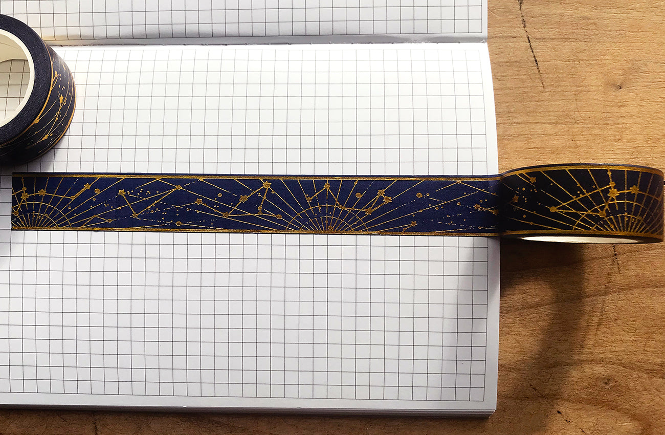 Gold foil constellation washi tape over gridded journal page on wooden table.