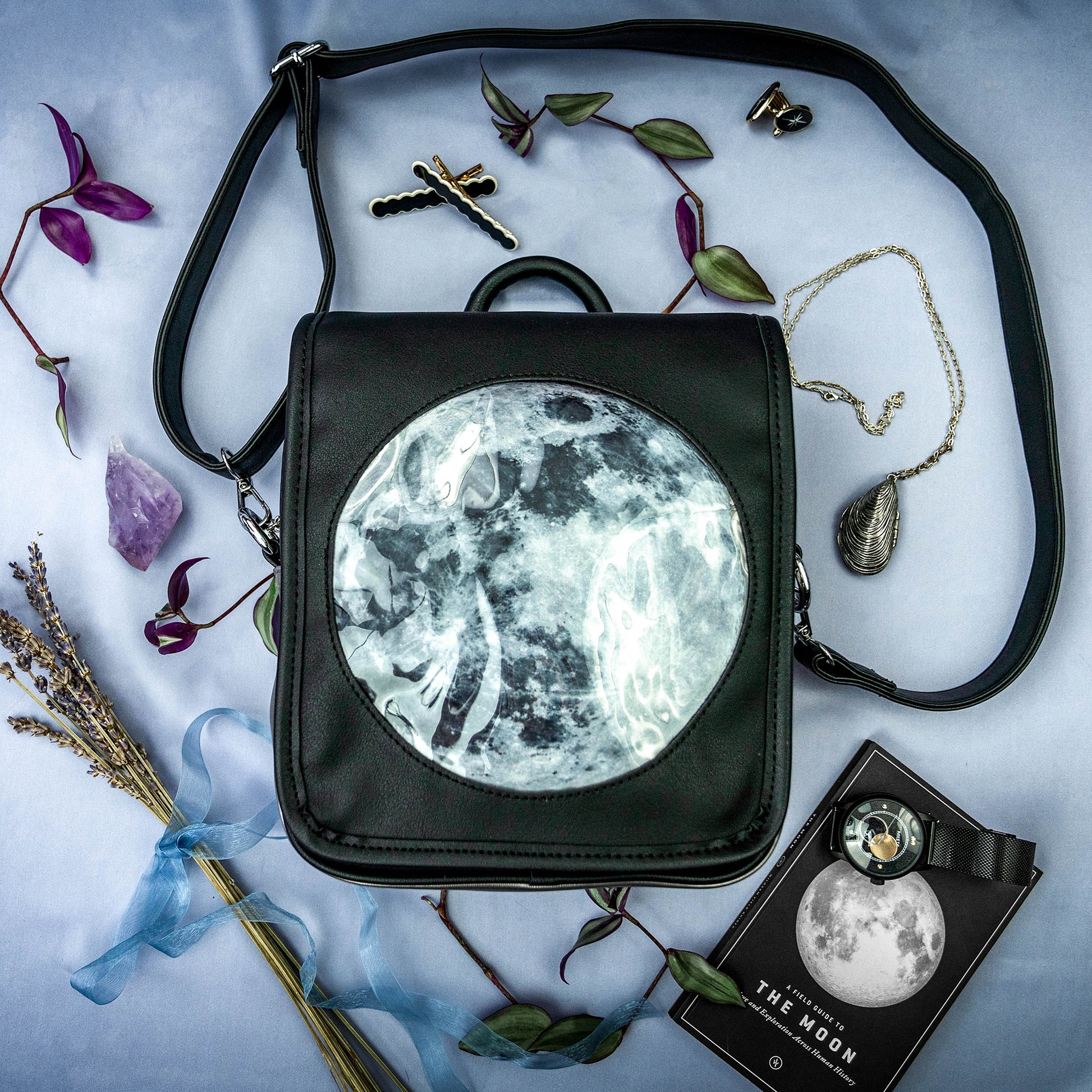 A flat lay photograph of the Moon Ita Bag on a smoky blue cloth amongst a collection of rocks, jewelry, greenery, ribbons, and thematic lunar objects.