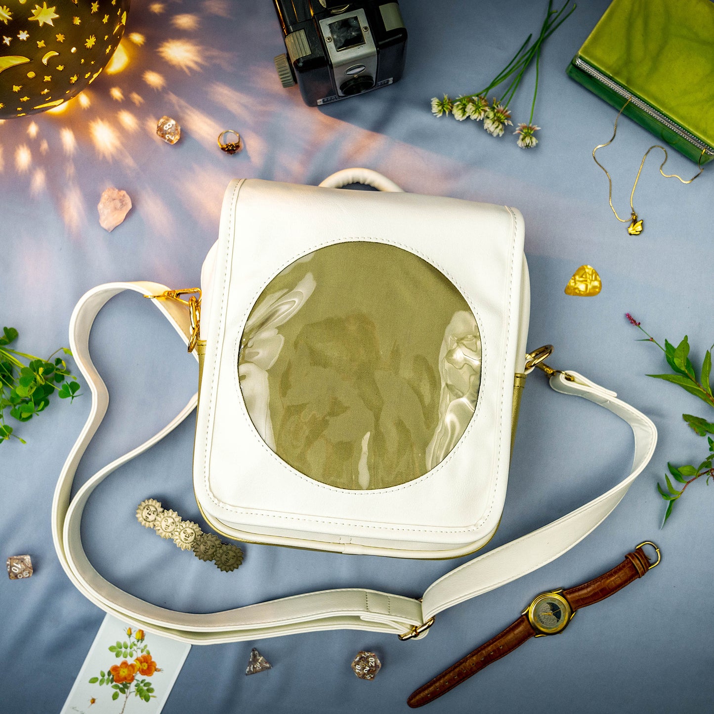 A flat lay photograph of the Sun Ita Bag on a sky blue cloth amongst a collection of rocks, sun-themed jewelry, yellow dice, a glowing constellation lamp, and greenery.