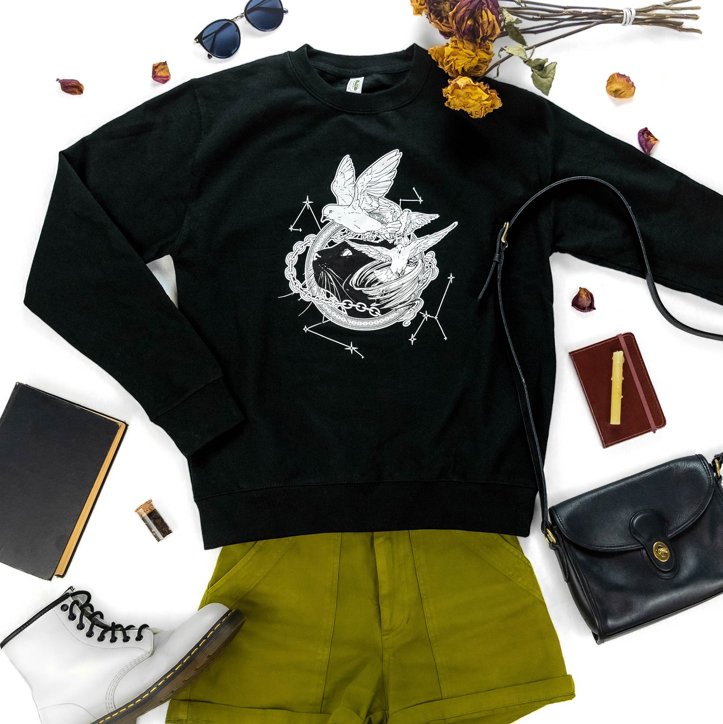 A flat lay photograph of the Tamora Pierce: Faithful and the Dust Spinners Sweatshirt in black, paired with various accessories, shorts, and floral elements.