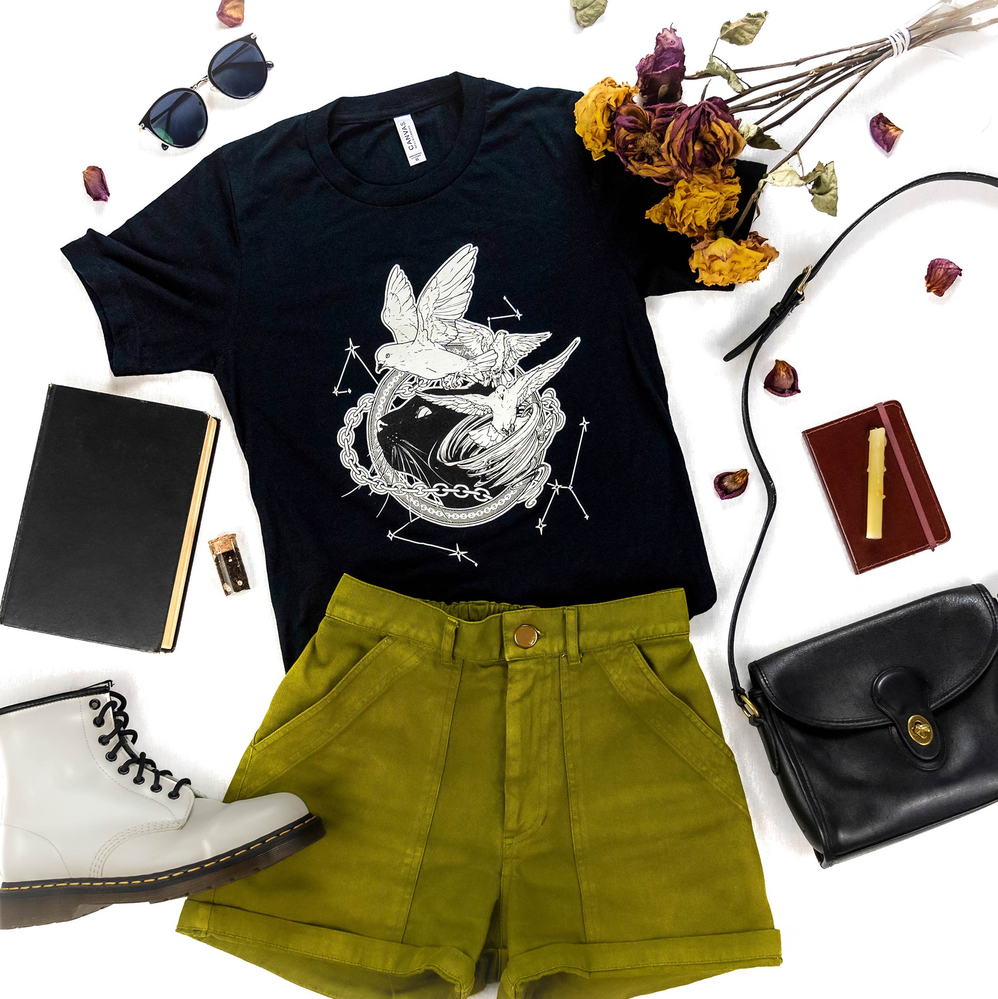 A flat lay photograph of the Tamora Pierce: Faithful and the Dust Spinners Tee in black, paired with various accessories, shorts, and floral elements.