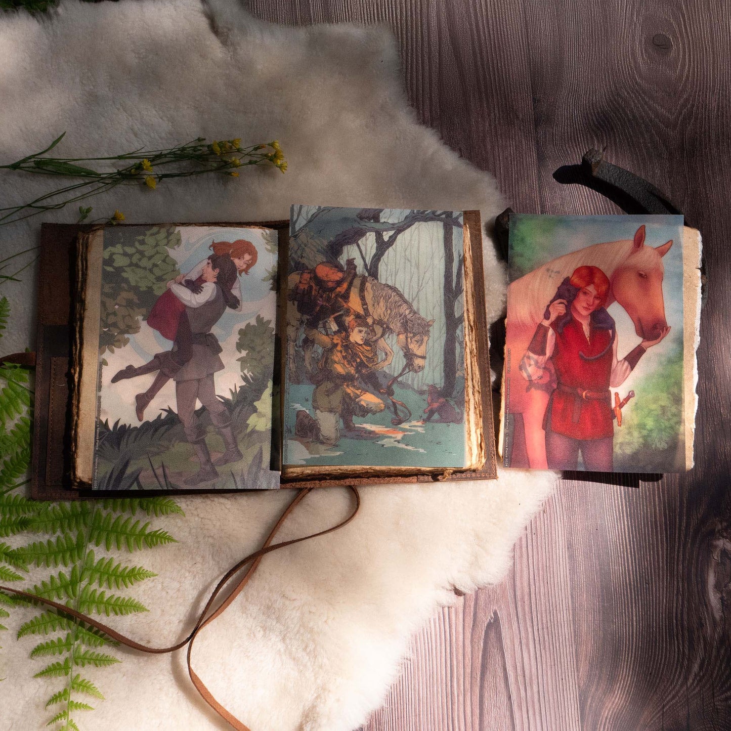 A photo of two of the page overlays placed inside a leatherbound journal, with the third page overlay resting beside the journal on top of a horseshoe. Ferns, sheepskin, and flowers adorn the display.