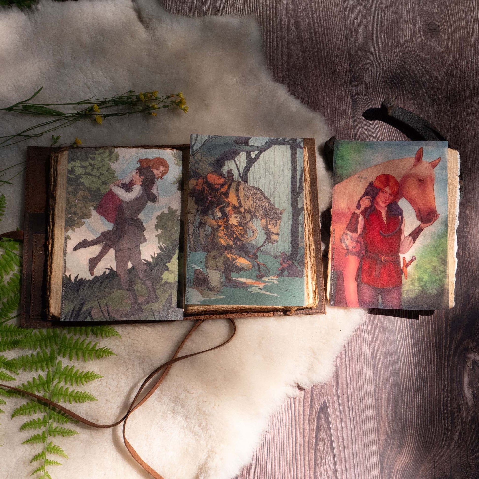 A photo of two of the page overlays placed inside a leatherbound journal, with the third page overlay resting beside the journal on top of a horseshoe. Ferns, sheepskin, and flowers adorn the display.