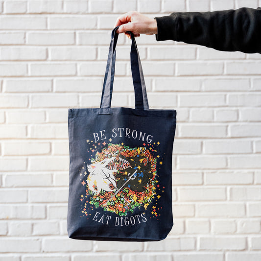 A model’s hand holding the Eat the Bigots Tote in front of a white brick wall.