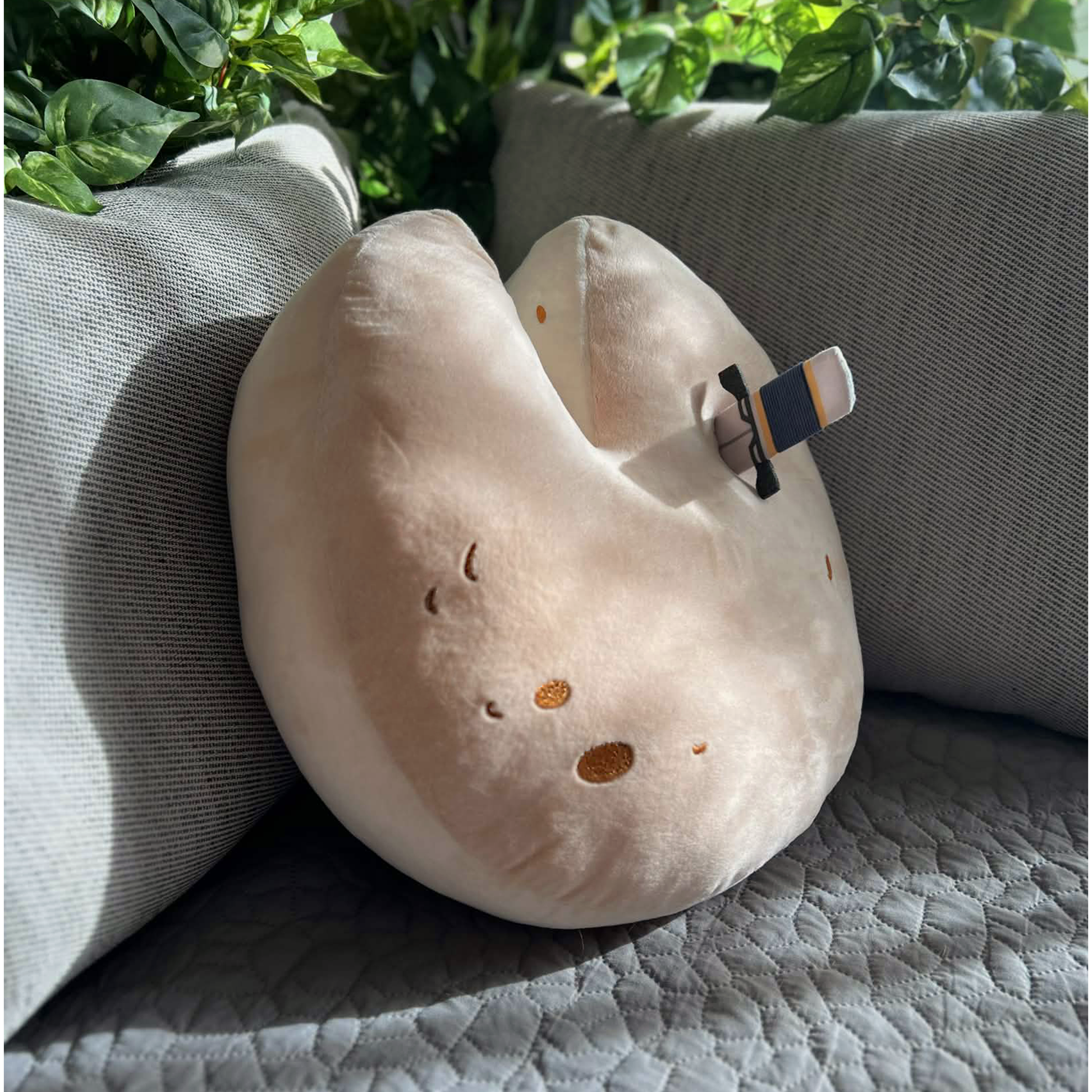 The Wedge of Destiny Shield Pillow is leaning face up against a grey couch at a three quarter angle. The pillow is round with a V cut at the top, as though someone has taken a slice of cheese. The pillow is a light tan color that ombres slightly darker on the front, with dark brown embroidered details to represent holes in the cheese.  A removable thin plush dagger is sticking out of the top right side of the pillow.