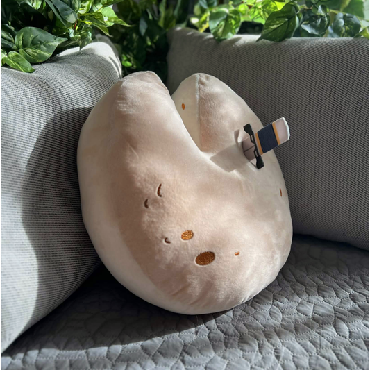 The Wedge of Destiny Shield Pillow is leaning face up against a grey couch at a three quarter angle. The pillow is round with a V cut at the top, as though someone has taken a slice of cheese. The pillow is a light tan color that ombres slightly darker on the front, with dark brown embroidered details to represent holes in the cheese.  A removable thin plush dagger is sticking out of the top right side of the pillow.