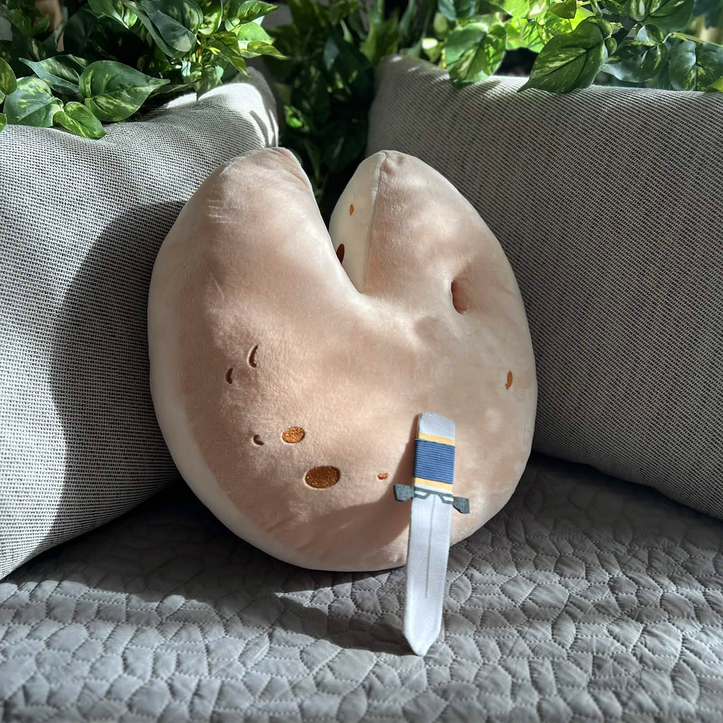 The Wedge of Destiny Shield Pillow is leaning face up against a grey couch at a three quarter angle.The removable dagger is removed from the pillow and is in turn resting against the shield. The dagger is light grey with a dark grey hilt and golden yellow details