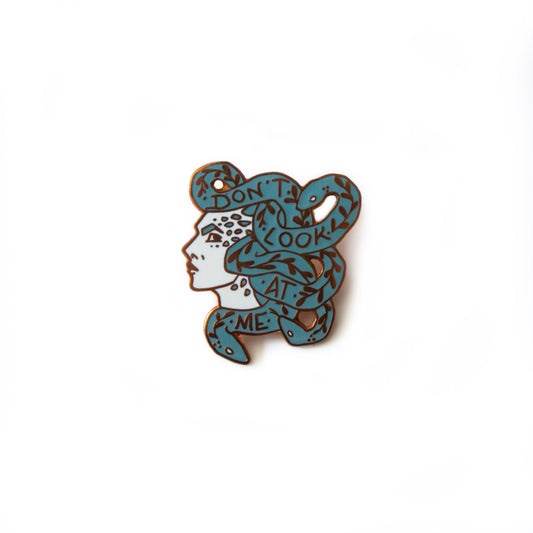 A product photo of an enamel pin depicting the face of Medusa in profile. Her skin is white with teal scales, and the snakes coiling around one another are teal with gold vines and leaves. Also in gold, the text on the snakes reads “Don’t Look At Me.”
