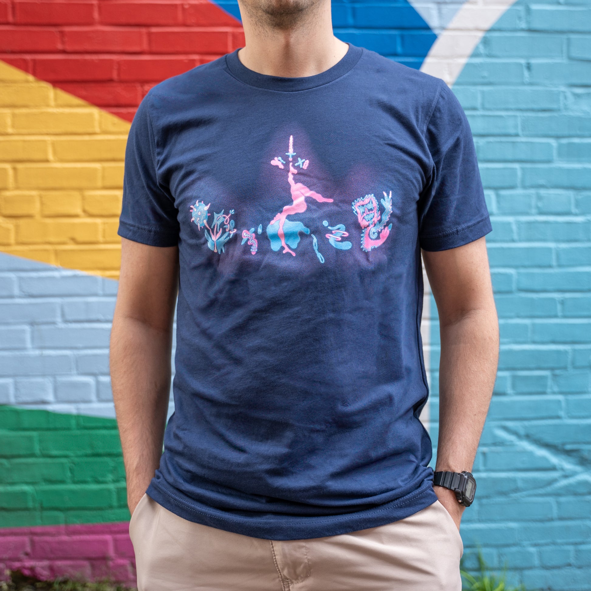 A model wearing the navy Pride Collaboration Tee in front of a colorfully painted brick wall, which makes the pastel pink and blues of the tee pop.