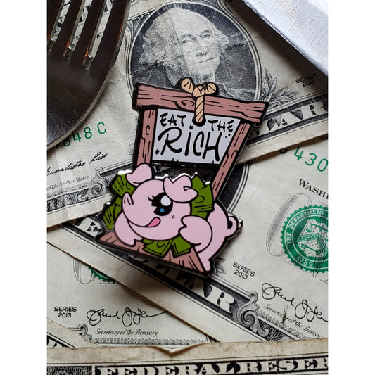 Black nickel enamel pin depicting light pink smiling pig in a guillotine whose blade reads "Eat the Rich." Pin is placed alongside fork and knife atop American dollar bills.
