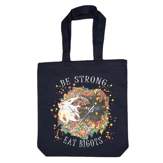  A flat lay photo of a black tote with a black wolf’s head and a white wolf’s head facing one another with their mouths open, flashing many sharp teeth. Two thin swords are crossing over each wolf. Surrounding them both is a pastel rainbow assortment of sparkles and flowers. The top arcing text reads “BE STRONG.” The bottom arcing text reads “EAT BIGOTS.”