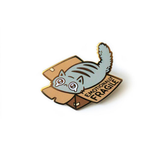 A product photo of an enamel pin that shows a fluffy grey cat stuffed into a barely taped together open brown box. The cat’s eyes are burbling with tears. The outer flap of the box reads “Emotionally Fragile.”