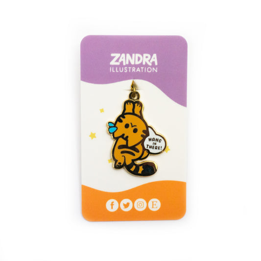 The Hang in There Zipper Pull on its purple, white, and orange wavy colorblocked backing card. The backing card says Zandra Illustration at the top in white, and displays icons that signify the artist is on facebook, twitter, instagram, and etsy along the bottom of the card.