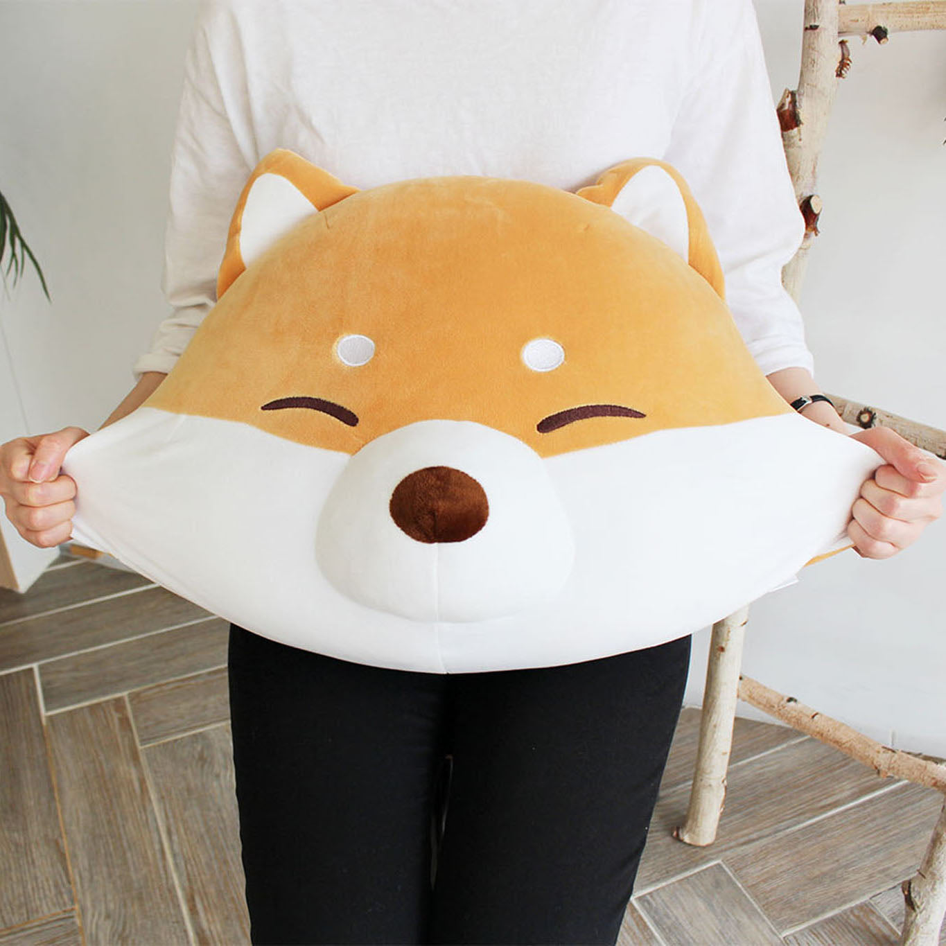 Front view of model stretching the Shiba cushion's cheeks with plants in the background.