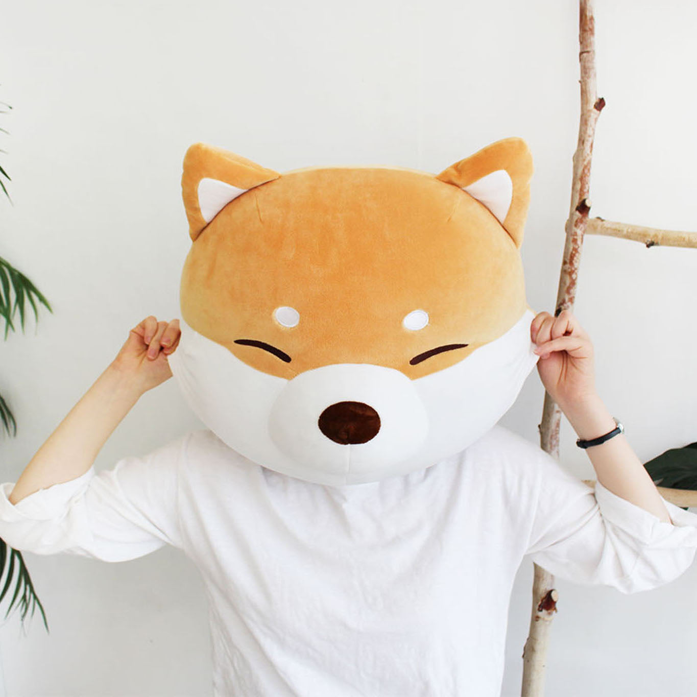 Model is holding the shiba face cushion in front of their face beside a birch ladder and a palm plant.